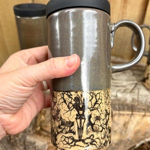 May include: A tall ceramic travel mug with a smooth, glossy dark gray upper section and a matte cream lower section featuring a detailed black ink illustration of a skeleton standing among tangled tree branches and various floral elements.