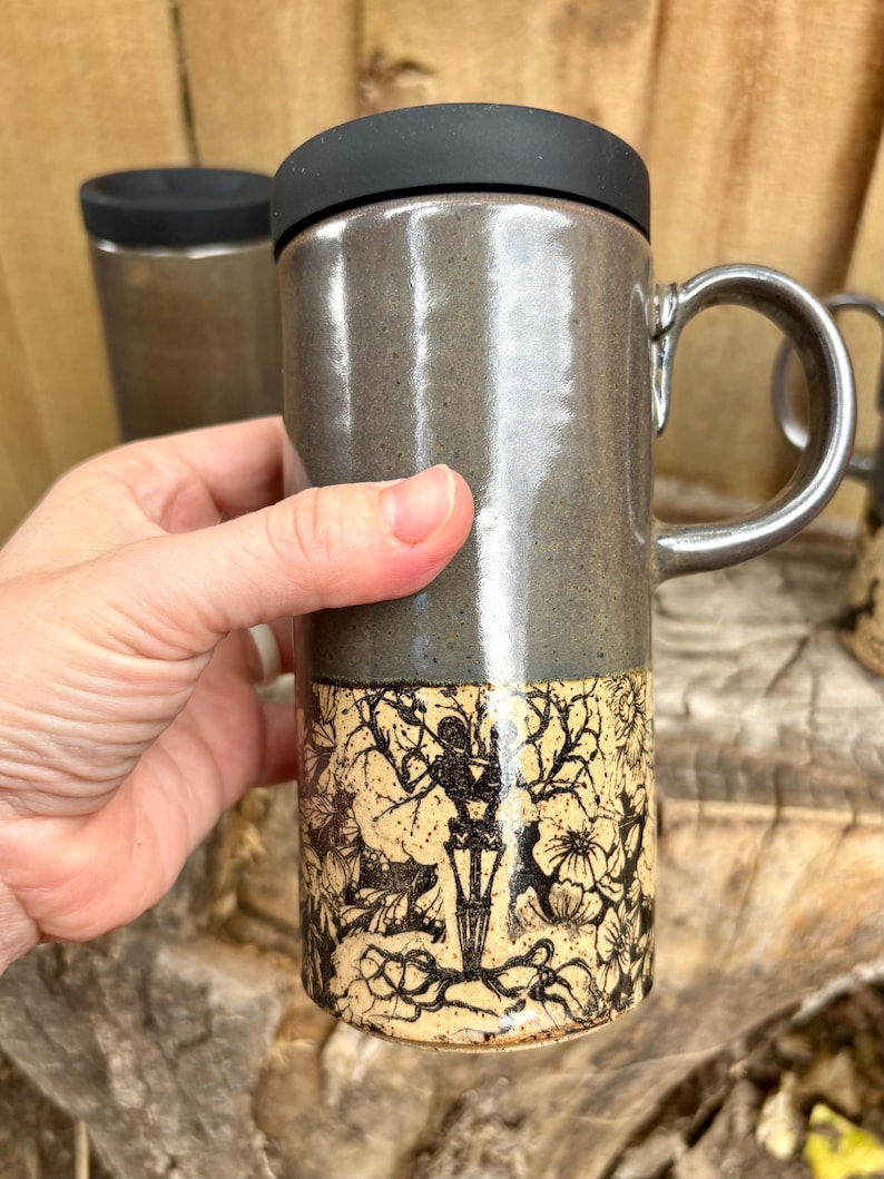 May include: A tall ceramic travel mug with a smooth, glossy dark gray upper section and a matte cream lower section featuring a detailed black ink illustration of a skeleton standing among tangled tree branches and various floral elements.