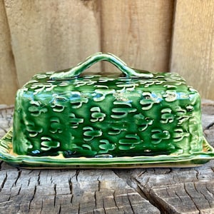 May include: A rectangular green ceramic butter dish with a glossy finish and a raised, repeating cactus pattern across the lid. The lid features a central arched handle, and the base has a matching green rim with a slightly scalloped edge.