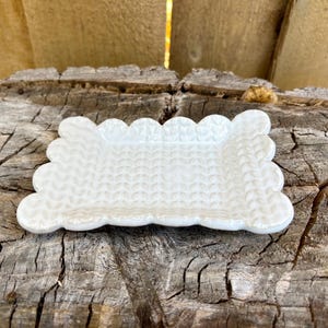 May include: A small, white ceramic dish with a scalloped edge and a textured surface. The rectangular dish is designed with a knitted pattern and is suitable for holding small items. The dish is displayed on a weathered wooden surface.