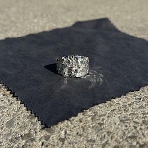 Könnte beinhalten: Ein silberner Ring mit einer strukturierten, unregelmäßigen Oberfläche. Der Ring liegt auf einem dunkelgrauen Tuch vor einem Betonhintergrund. Das Design des Rings hat ein natürliches, organisches Aussehen.