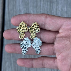 May include: Four stud earrings in the image. Two are gold-colored, and two are silver-colored. Each earring has a textured, irregular shape, resembling a nugget or organic form. The earrings are displayed against a neutral background.