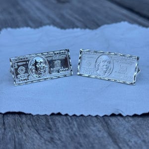 May include: Two silver-toned cufflinks, each designed to resemble a U.S. $100 bill. The cufflinks feature detailed engravings of the bill's design, including the portrait of Benjamin Franklin and the number 100.