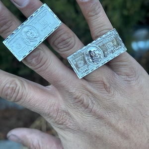 May include: Two silver rings, each shaped like a U.S. one-hundred-dollar bill, are displayed on fingers. The rings feature detailed engravings of the bill's design, including the portrait and text.