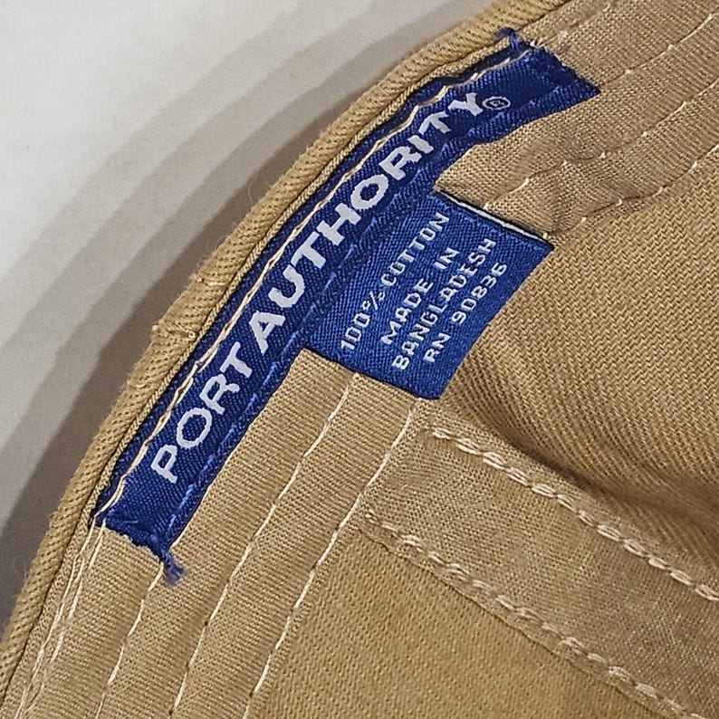 May include: Close-up of a tan baseball cap with a blue label. The label reads "PORT AUTHORITY" in white text. Below the brand name, the label states "100% COTTON MADE IN BANGLADESH RN 90836". The cap has white stitching.