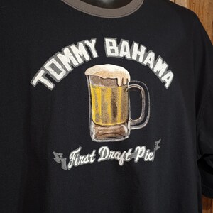 May include: A black t-shirt featuring a graphic of a beer mug filled with yellow liquid and white foam. The text TOMMY BAHAMA is arched above the mug, and the text First Draft Pick is printed on a banner below the mug in white lettering.