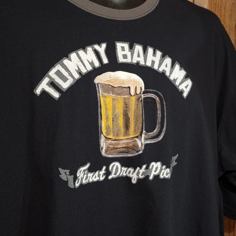 May include: A black t-shirt featuring a graphic of a beer mug filled with yellow liquid and white foam. The text TOMMY BAHAMA is arched above the mug, and the text First Draft Pick is printed on a banner below the mug in white lettering.