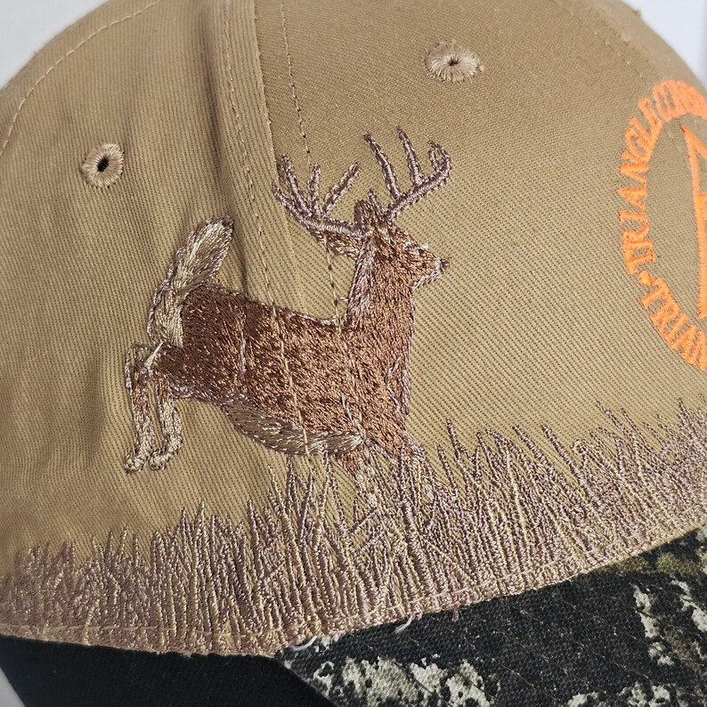 May include: Tan baseball cap with an embroidered brown deer leaping over tall grass. The cap features a camouflage brim and the text "TRIANGLE" in orange. A classic accessory for outdoor enthusiasts.
