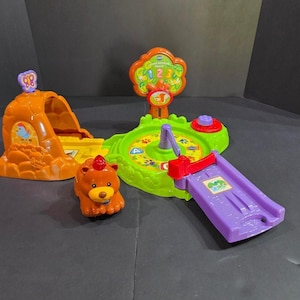 May include: A VTech Forest Adventure playset featuring an orange cave with a purple butterfly, a green circular base with a central rotating arm, a red button, and a purple slide. A small brown bear figure with a red light on its head is included.