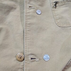 May include: Close-up of a tan jacket with visible stitching and a pocket with a zipper. The jacket has two buttons and two silver coins are visible. The fabric appears to be a durable, possibly canvas material.
