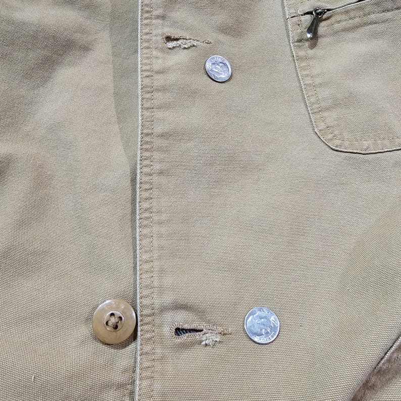 May include: Close-up of a tan jacket with visible stitching and a pocket with a zipper. The jacket has two buttons and two silver coins are visible. The fabric appears to be a durable, possibly canvas material.