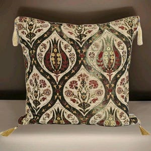 May include: Square decorative pillow with a floral pattern in shades of cream, burgundy, and olive green. The pillow has a dark green border and gold tassels on each corner. The pattern features stylized flowers and leaves.