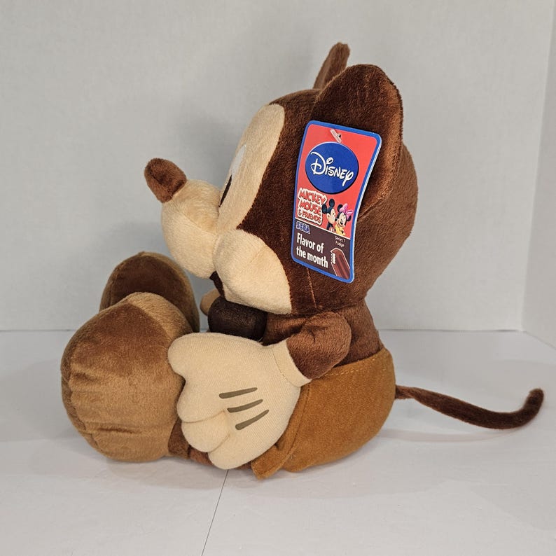 Puede incluir: Un peluche de un personaje de dibujos animados, principalmente marr&oacute;n y beige, con orejas grandes y una cola larga. El personaje tiene guantes blancos y una etiqueta que dice "Disney Mickey Mouse & Friends Flavor of the month". El juguete est&aacute; sentado.