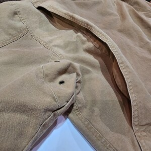 May include: Close-up of a tan work jacket, highlighting the fabric's texture and stitching. The jacket has a pocket and a metal grommet. The design suggests durability and utility, typical of workwear.