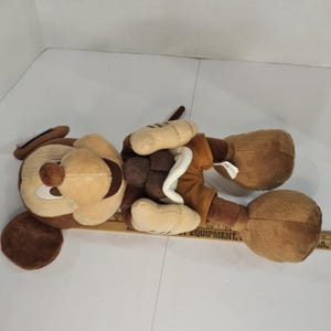 Puede incluir: Un peluche de perro en tonos marr&oacute;n, beige y crema. El juguete tiene orejas grandes y ca&iacute;das, un hocico redondo y un collar blanco. El perro est&aacute; tumbado boca arriba, con las patas en el aire. El juguete es suave y tierno.