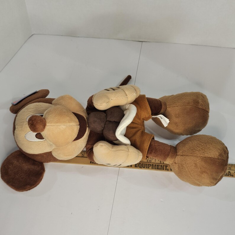 Puede incluir: Un peluche de perro en tonos marr&oacute;n, beige y crema. El juguete tiene orejas grandes y ca&iacute;das, un hocico redondo y un collar blanco. El perro est&aacute; tumbado boca arriba, con las patas en el aire. El juguete es suave y tierno.