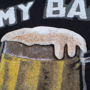 May include: A black t-shirt featuring a graphic of a beer mug filled with golden liquid and topped with thick white foam. The text MY BA is printed in white block letters above the beer mug graphic, which has a textured, raised appearance.