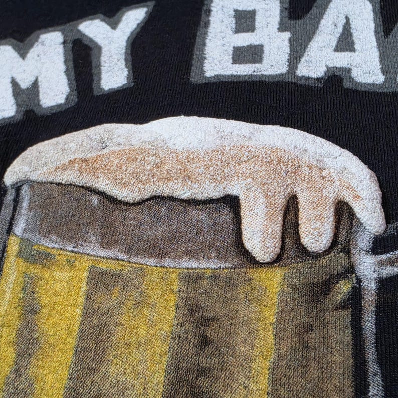 May include: A black t-shirt featuring a graphic of a beer mug filled with golden liquid and topped with thick white foam. The text MY BA is printed in white block letters above the beer mug graphic, which has a textured, raised appearance.