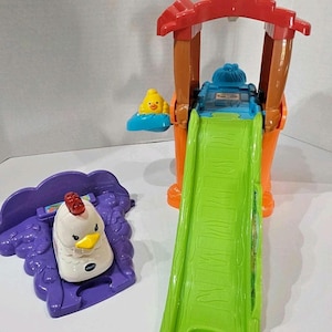 May include: A colorful VTech farm playset featuring a chicken and a slide. The playset includes a white chicken with a red comb, a green slide, and a red and brown structure. The toy is designed for young children.