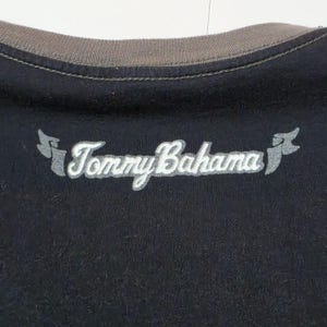 May include: A black shirt featuring a brown ribbed crew neckline. The Tommy Bahama brand name is embroidered in white script across the chest, flanked by two small, stylized grey flag-like icons on either side of the text.