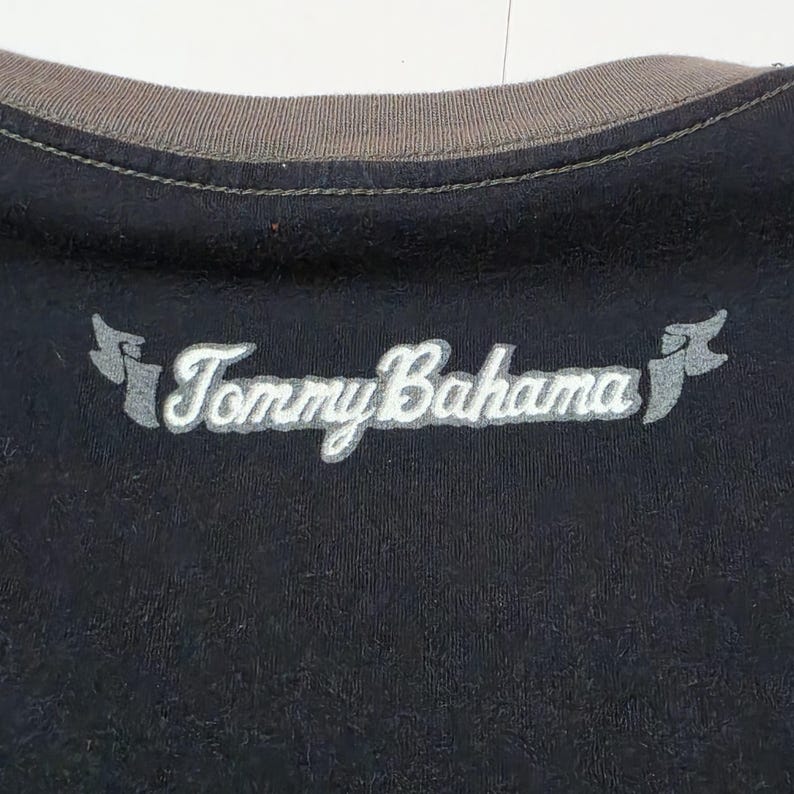 May include: A black shirt featuring a brown ribbed crew neckline. The Tommy Bahama brand name is embroidered in white script across the chest, flanked by two small, stylized grey flag-like icons on either side of the text.