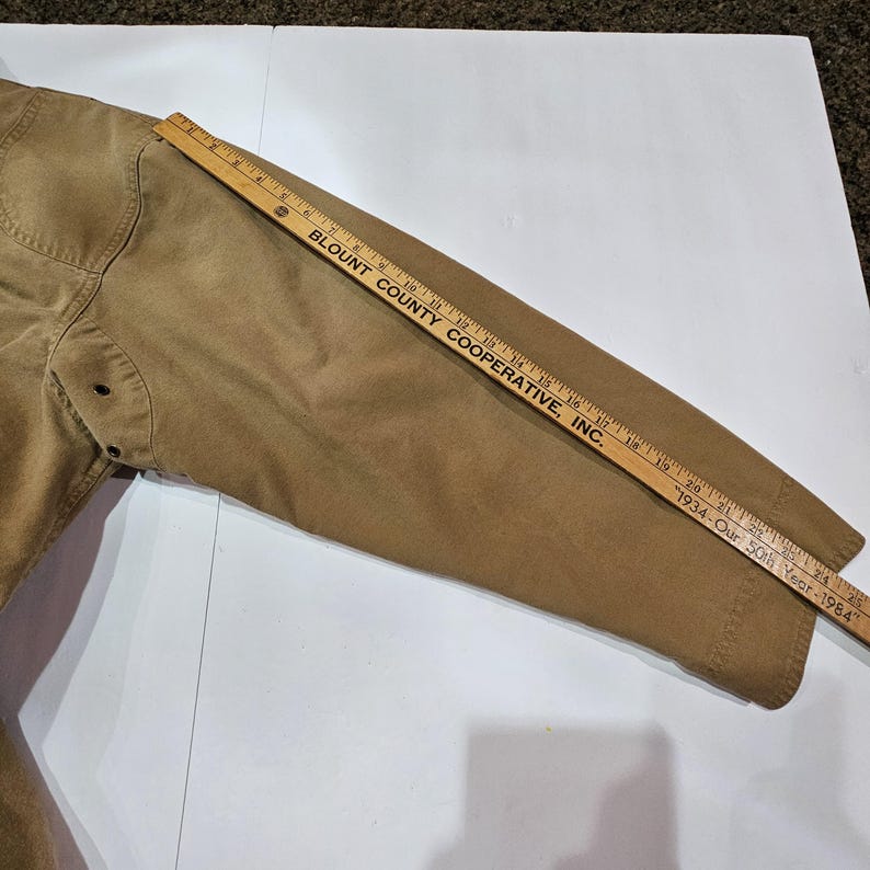 May include: A tan jacket sleeve with visible stitching and button details. A wooden ruler with the text "BLOUNT COUNTY COOPERATIVE, INC." and "1934-Our 50th Year-1984" is placed across the sleeve.