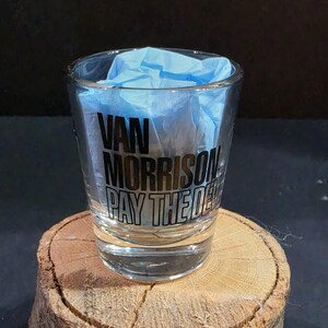 May include: A clear glass shot glass featuring black printed text that reads Van Morrison Pay The Devil. The interior of the glass is filled with light blue tissue paper, and the glass has a thick, sturdy base with a smooth, transparent finish.