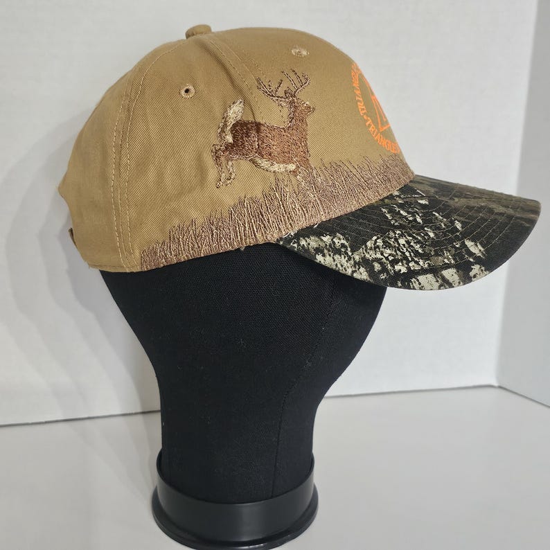 May include: Tan baseball cap with a brown embroidered deer leaping over a patch of grass. The bill of the cap has a camouflage pattern. The cap also has the text "Whitetail Triangle" in orange.