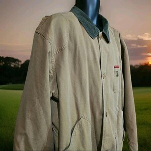 May include: A tan corduroy jacket with a dark green collar. The jacket has two large front pockets and a small red and white logo on the chest. The jacket is buttoned up and displayed on a mannequin.