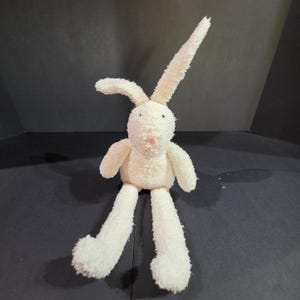 May include: A plush stuffed rabbit toy with cream-colored, textured fabric. The rabbit features one long upright ear and one folded ear, a small pink stitched nose, and two dark eyes. It has long arms and long legs extending from a round body.