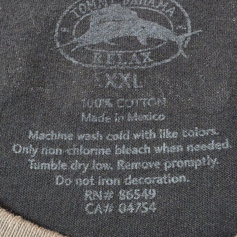 May include: A black fabric garment tag featuring the Tommy Bahama Relax logo with a marlin graphic, size XXL, and 100% cotton composition. The text includes care instructions such as machine wash cold, tumble dry low, and do not iron decoration.