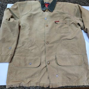 May include: A tan jacket with a green collar and multiple pockets. The jacket has a button-up front and silver buttons. It features a small chest pocket and two large front pockets. The jacket is made of a durable material, suitable for outdoor wear.