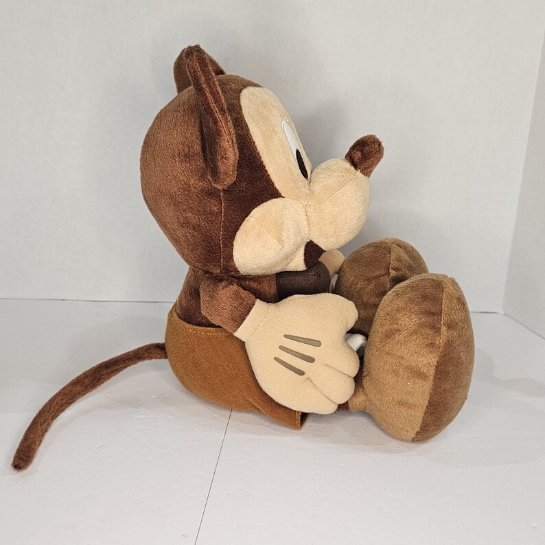 Puede incluir: Un peluche de un rat&oacute;n de dibujos animados en posici&oacute;n sentada. El juguete presenta un cuerpo, orejas y cola marrones, con cara, manos y pies de color beige. El rat&oacute;n lleva pantalones cortos marrones. El juguete est&aacute; dise&ntilde;ado para parecerse a un personaje de dibujos animados cl&aacute;sico.