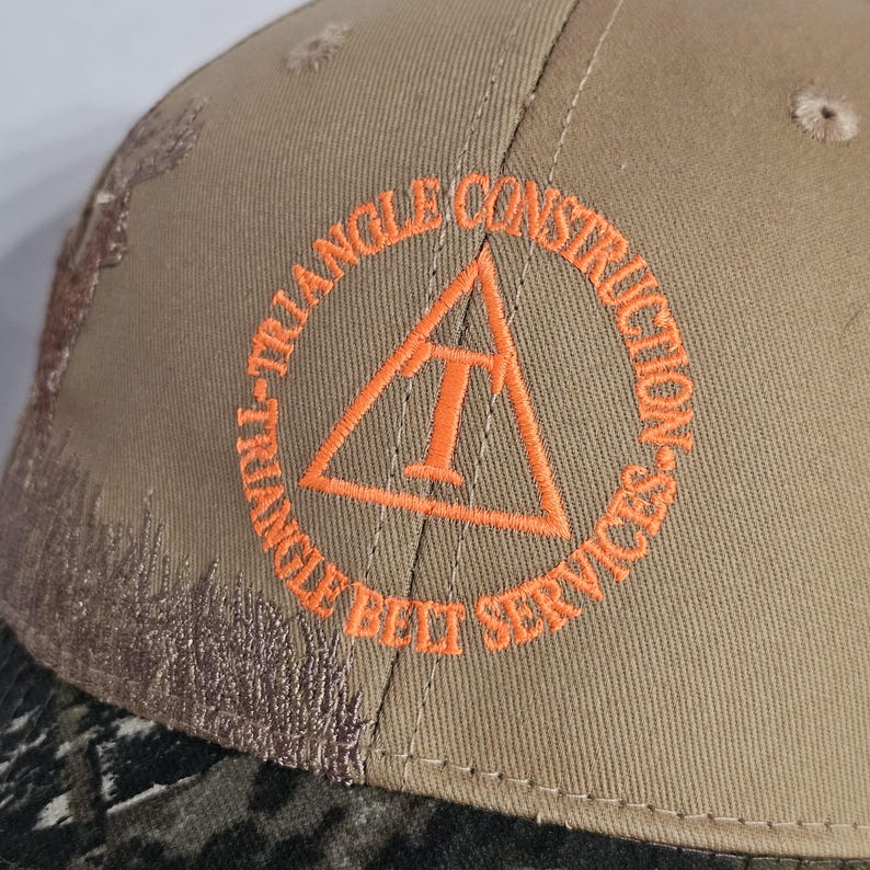 May include: A tan baseball cap with the embroidered text "TRIANGLE CONSTRUCTION BELT SERVICES" in orange. The cap features a camouflage pattern on the brim and a small embroidered design on the side.