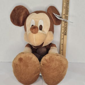 Puede incluir: Un peluche de Mickey Mouse en tonos marrones y beige. El peluche tiene orejas grandes y redondas, una cara sonriente y pies de gran tama&ntilde;o. Una regla de madera est&aacute; al lado del juguete.
