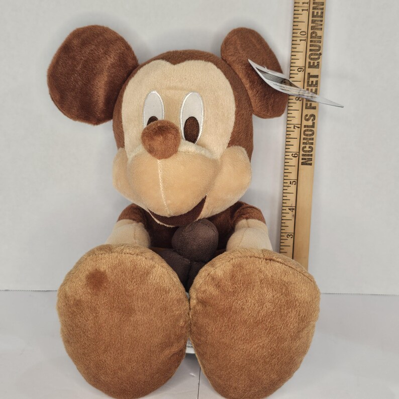 Puede incluir: Un peluche de Mickey Mouse en tonos marrones y beige. El peluche tiene orejas grandes y redondas, una cara sonriente y pies de gran tama&ntilde;o. Una regla de madera est&aacute; al lado del juguete.