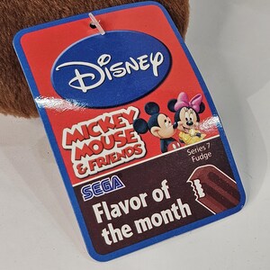 Puede incluir: Una etiqueta rectangular de Disney Mickey Mouse & Friends con fondo rojo y borde azul. La etiqueta presenta a Mickey y Minnie Mouse, el texto "Flavor of the month" y "Series 7 Fudge".