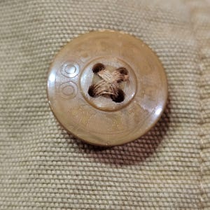 May include: Close-up of a beige button with the word "STC" and other text around the edge. The button has a cross-stitched centre and is attached to beige fabric.