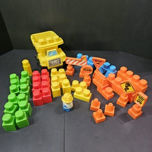 May include: A yellow Tonka dump truck toy with a set of colorful building blocks and construction-themed accessories. The set includes orange, green, red, and yellow blocks, along with road signs and a construction worker figurine. The word "SLOW" is visible on a sign.