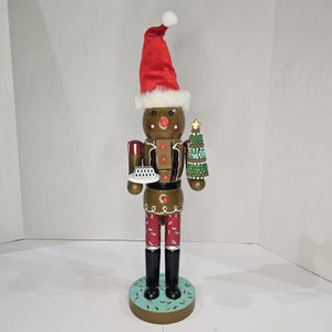 May include: A decorative gingerbread man nutcracker figurine. The figure wears a red Santa hat with white trim, red pants, and black boots. It holds a plate and a small Christmas tree. The base is light blue with sprinkles.