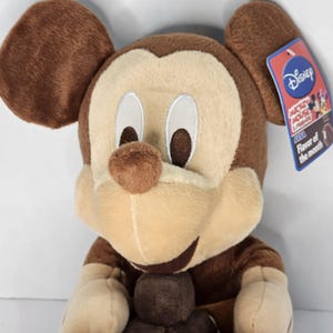 Puede incluir: Un peluche de Mickey Mouse en tonos marr&oacute;n y beige. El juguete tiene orejas grandes, una nariz redonda y una cara sonriente. Una etiqueta de Disney est&aacute; adjunta, con el texto "Mickey Mouse & Friends" y "Flavor of the month".