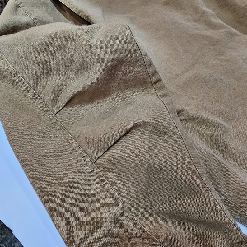 May include: Close-up of tan trousers. The fabric looks like a robust cotton or blend, with visible stitching along the seams. The trousers are folded, showing the details of the pockets and the overall construction. The colour is a neutral, versatile shade.