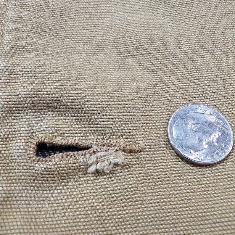 May include: Close-up of tan fabric with a buttonhole and a coin. The buttonhole is frayed, showing the texture of the fabric. The coin is silver.