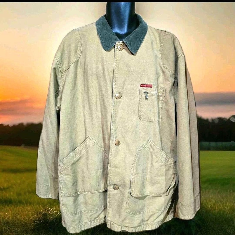 May include: Tan jacket with a dark green corduroy collar, button closure, and multiple pockets. Features a small red and white label on the chest pocket. The jacket has a classic, utilitarian design.