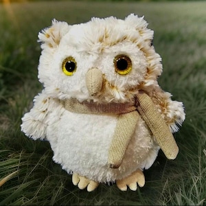 May include: A plush owl toy with a cream-colored body and a tan scarf. The owl has large, yellow eyes and a tan beak. The soft toy is sitting on a bed of green grass.
