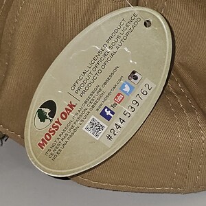 May include: A tan baseball cap with a tag attached. The tag features the Mossy Oak logo and text in English, French, and Spanish, along with social media icons and a product number.