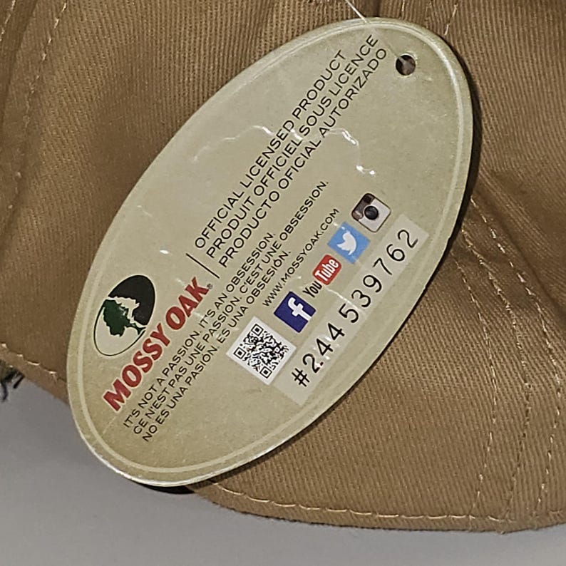 May include: A tan baseball cap with a tag attached. The tag features the Mossy Oak logo and text in English, French, and Spanish, along with social media icons and a product number.