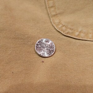 May include: A silver quarter coin featuring an eagle design, placed on a tan fabric. The coin's details are clear, and the fabric has a textured look. The coin is approximately 2.4 cm in diameter.