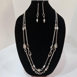 May include: A silver-toned necklace and earring set displayed on a black mannequin. The necklace features two strands with various beads in shades of white, brown, and gray. The earrings are long, dangling, and have matching beads. The jewelry is designed with a mix of chain and bead elements.