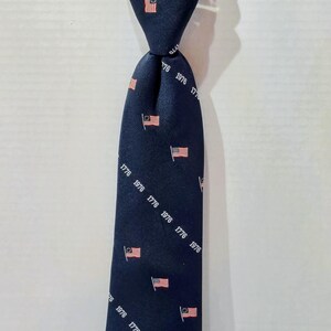 May include: Navy blue zipper tie with a repeating pattern of small American flags and the year "1776". The tie has a pre-tied knot and is attached to a cardboard tag that reads "ZIPPER TIES".